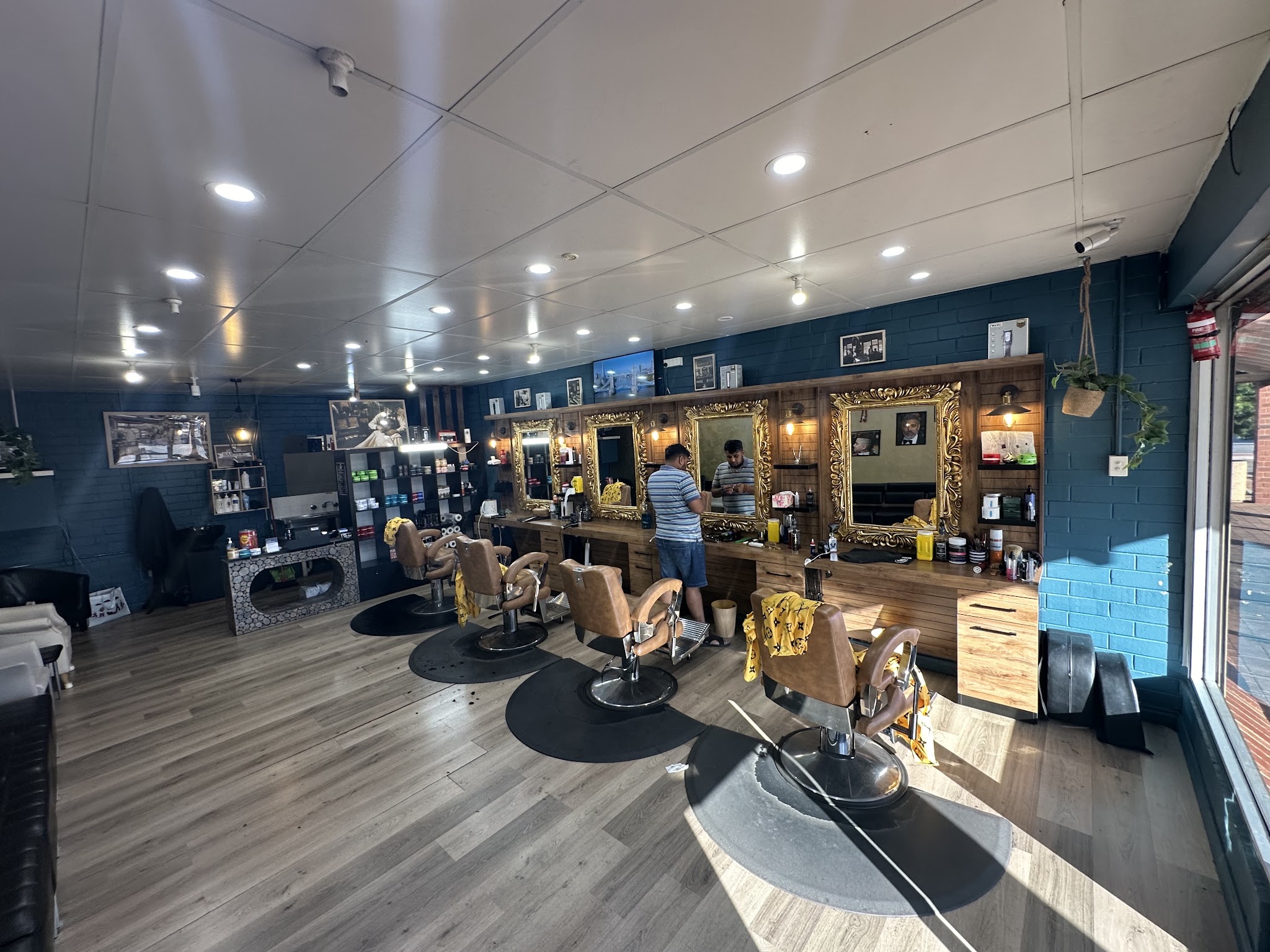Daylight inside the shop: tan leather barber chairs, gold mirrors along a blue brick wall and a barber at work